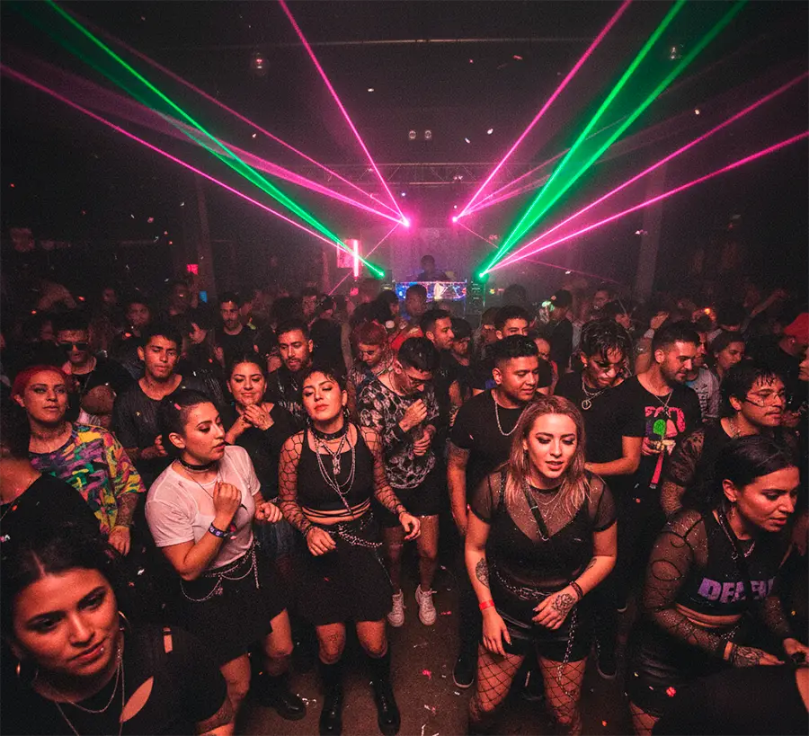 Público joven y diverso en una fiesta techno underground en Santiago, bailando bajo intensas luces láser de color rosa y verde.